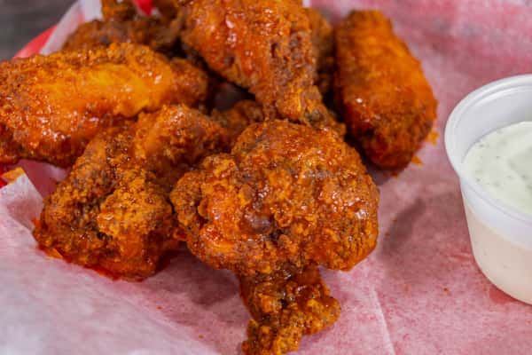 Crispy buffalo-style chicken wings coated in spicy sauce, served in a red basket with pink parchment paper and a side of creamy ranch dressing.