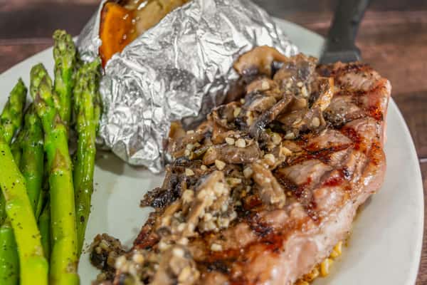 Grilled steak topped with sautéed mushrooms and garlic, served with a baked sweet potato in foil and a side of seasoned asparagus on a white plate.
