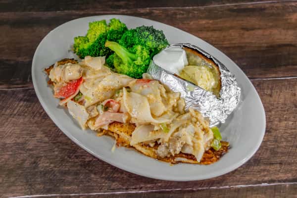 Blackened fish fillet topped with creamy seafood and vegetable medley, served with steamed broccoli and a foil-wrapped baked potato topped with butter and sour cream, showcasing a hearty coastal comfort dish.