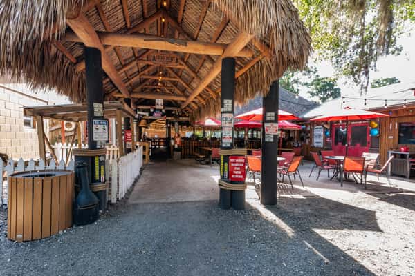 Thatched-roof tiki walkway leading to Cherry Pocket's vibrant outdoor patio dining area with red umbrellas and rustic seating under the Florida sun.