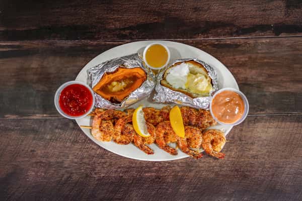 Grilled shrimp skewers served with a baked potato, sweet potato, cocktail sauce, and drawn butter, plated for a hearty coastal seafood entrée.