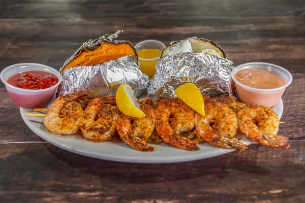 Grilled shrimp skewers seasoned with Cajun spices, served with baked sweet and russet potatoes wrapped in foil, lemon wedges, and a trio of dipping sauces at a rustic seafood restaurant.