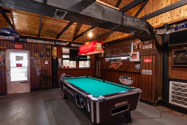 Cozy game room with a classic bar-style pool table under a red Budweiser light, surrounded by vintage beer signs, license plates, and wood-paneled walls, creating a nostalgic dive bar atmosphere perfect for casual hangouts.