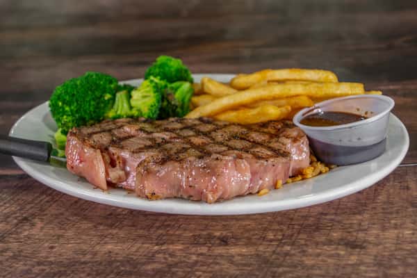 Close-up of grilled steak entrée with golden fries, broccoli, and a cup of brown dipping sauce, highlighting a hearty steakhouse-style meal.