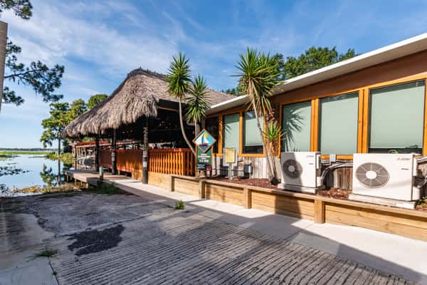 Thatched-roof tiki bar and restaurant beside a serene lakeshore, framed by tropical landscaping and wooden siding, showcasing waterfront dining at Cherry Pocket in Florida.