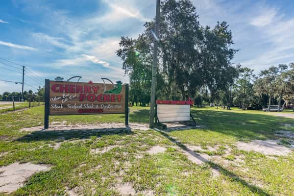 Roadside sign for Cherry Pocket Steak, Seafood Shak & Oyster Bar set against grassy grounds and shaded by oak trees, welcoming guests to a laid-back Florida fish camp experience.