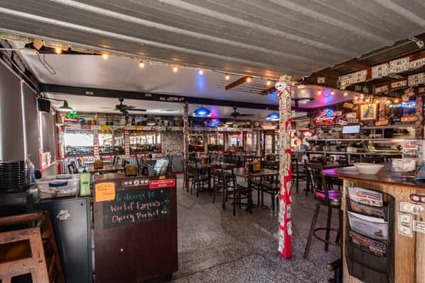 Wide-angle view of Cherry Pocket's lively indoor dining area with string lights, mismatched decor, and walls plastered in bills and memorabilia for a fun, dive-bar-style ambiance.