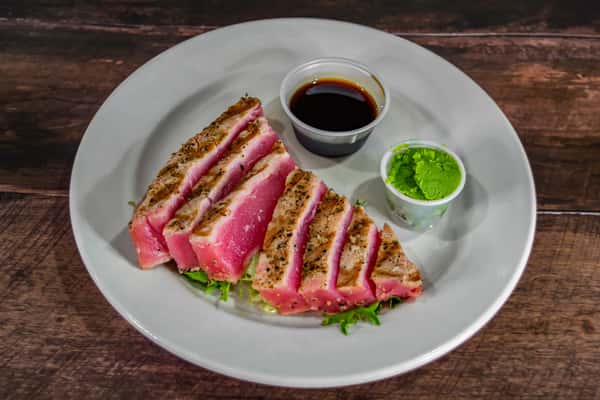 Sliced seared ahi tuna served rare, accompanied by wasabi and soy dipping sauce, presented over fresh greens on a white plate.