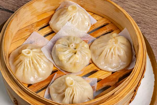 Five soup dumplings with pleated tops arranged in a bamboo steamer lined with parchment squares.