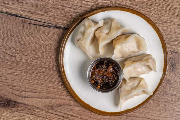 Six steamed dumplings arranged around a cup of soy-based dipping sauce on a white plate with a brown rim.