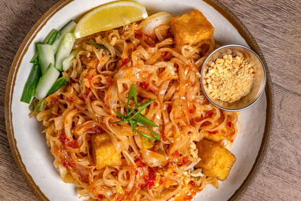 Tofu pad thai topped with scallions, served with crushed peanuts, cucumber sticks, and a lemon wedge on the side.