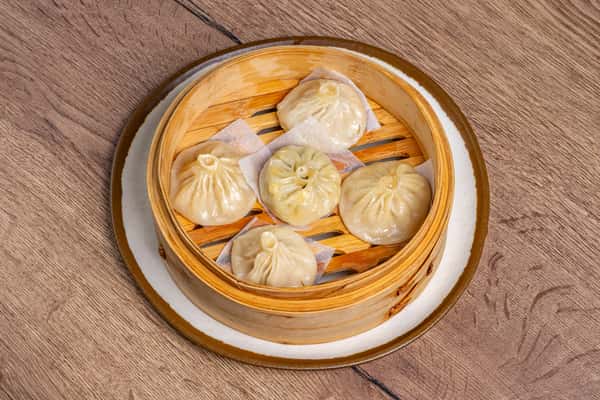 Five soup dumplings with pleated tops arranged in a bamboo steamer lined with parchment squares.