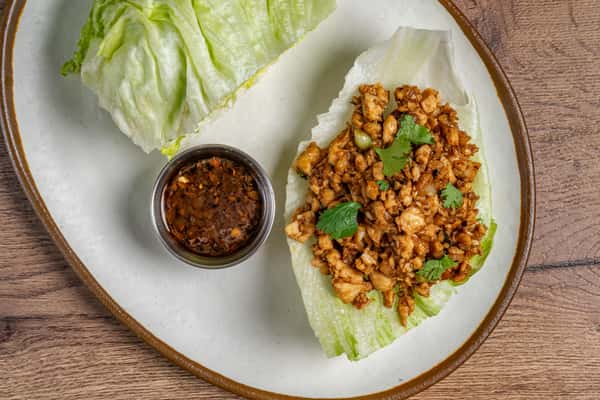 Minced chicken lettuce wrap topped with cilantro, served with a side of soy-based dipping sauce on a white plate.