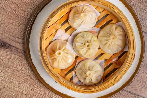 Five soup dumplings with pleated tops arranged in a bamboo steamer lined with parchment squares.
