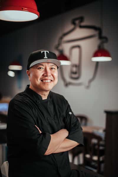 Smiling chef in a black uniform and baseball cap standing inside a modern restaurant with a rice cooker logo on the wall.