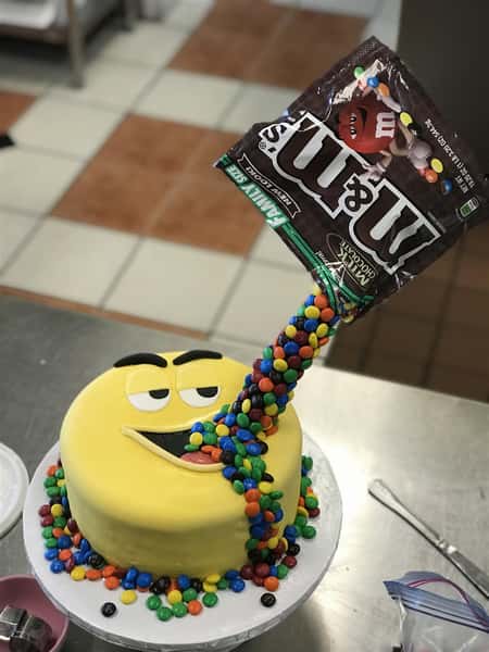 an M&M themed cake with a bag of M&Ms pouring into an M&M character's mouth