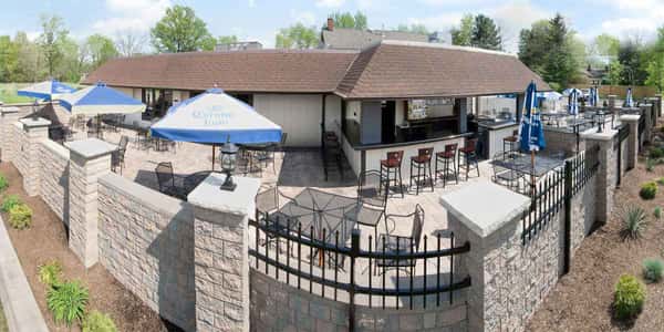 Outdoor patio bar with stone walls, umbrellas, and high-top seating surrounding a covered building on a sunny day.