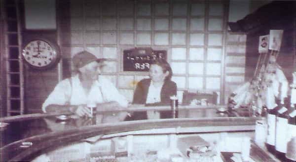 Vintage-style photo of two people seated at a curved bar counter with a wall clock and shoji-style panels behind them.