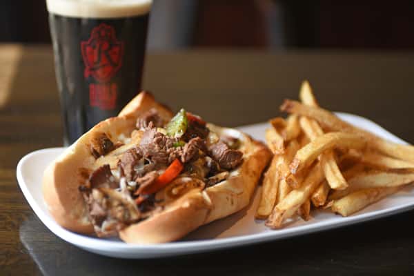 Philly cheesesteak with sautéed peppers and mushrooms, served with fries and a dark beer in background.