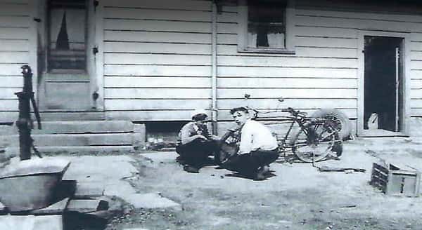 Black-and-white photo of two boys crouching beside a bicycle outside a wooden house with a hand water pump nearby.