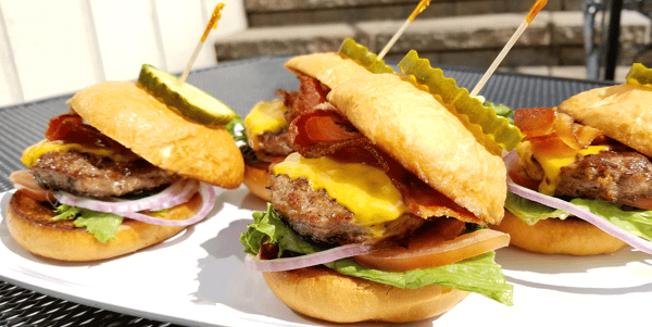 Three bacon cheeseburger sliders with lettuce, tomato, red onion, and pickles on toasted buns.