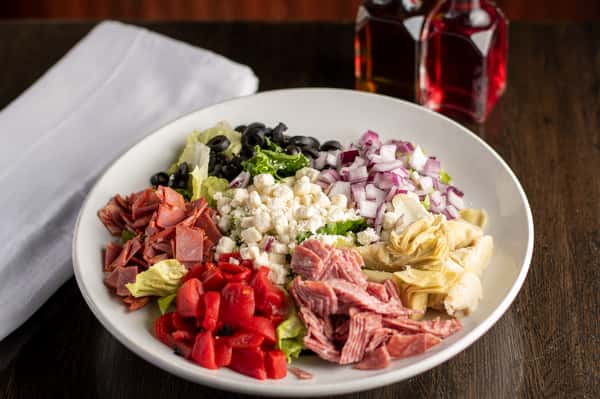 Antipasto salad with salami, ham, red peppers, olives, red onion, artichokes, and feta over lettuce in a white bowl.