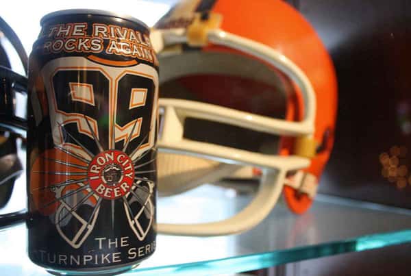 Iron City Beer can with "The Rivals Rocks Again 59" design beside an orange football helmet on a glass shelf.