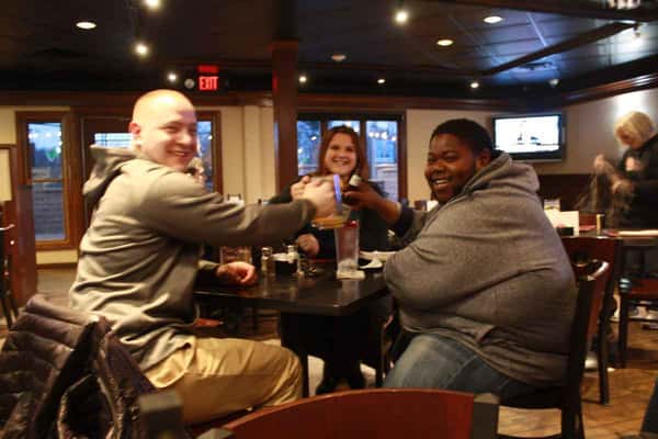 Three friends smiling and toasting drinks at a restaurant table, seated indoors under warm lighting.