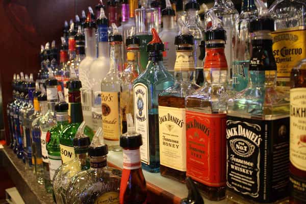 Liquor bottles lined up on a bar shelf, including whiskey, vodka, rum, and tequila with pour spouts attached.