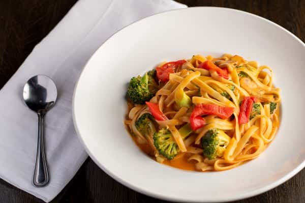 Creamy fettuccine pasta with broccoli and red bell peppers in a white bowl, served with a spoon and napkin on table.
