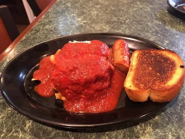 Hearty Italian-style meatloaf smothered in rich marinara sauce, served with a grilled sausage link and thick-cut toasted garlic bread on a black plate — a comforting and savory homestyle meal.