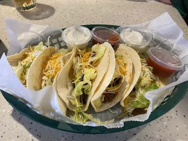 Trio of soft-shell tacos filled with seasoned meat, shredded lettuce, and cheese, accompanied by cups of sour cream and salsa — a vibrant Tex-Mex dish ideal for casual dining.