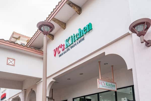 Exterior of VC's Kitchen, a casual eatery offering fresh salads, wraps, and more, located in a Mediterranean-style shopping plaza.