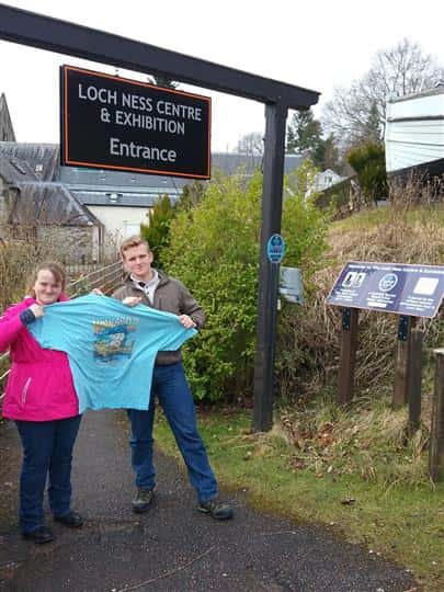 Man and woman holding Howard's Pub t-shirt in front of Loch Ness Centre entrance.