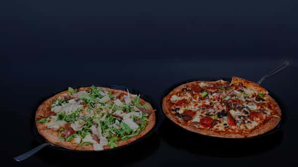 Two pizzas side by side—one topped with arugula and shaved parmesan, the other with assorted meats, olives, and vegetables.