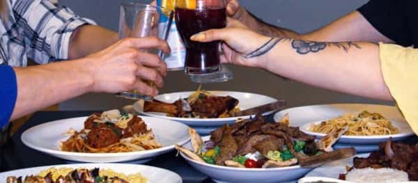 Four people clinking glasses over a table full of diverse pasta and meat dishes, celebrating with a shared meal.