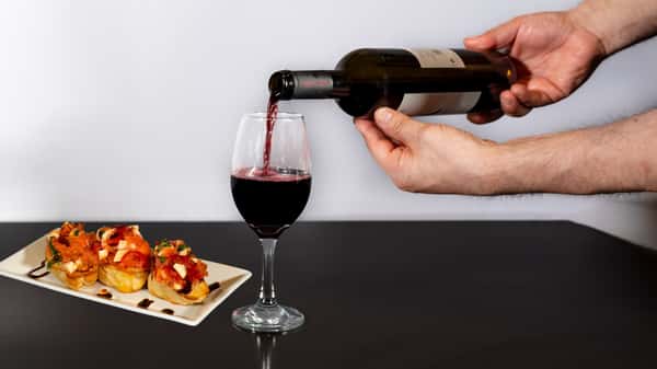 Red wine being poured into a glass next to a plate of bruschetta topped with tomatoes, mozzarella, and balsamic glaze.