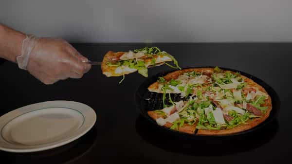 A gloved hand serves a slice of thin-crust pizza topped with arugula, shaved parmesan, and prosciutto onto a white plate.