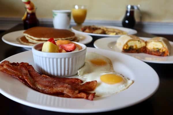 Breakfast plate with sunny-side-up eggs, crispy bacon, and fruit salad, with pancakes and breakfast burritos in the background.