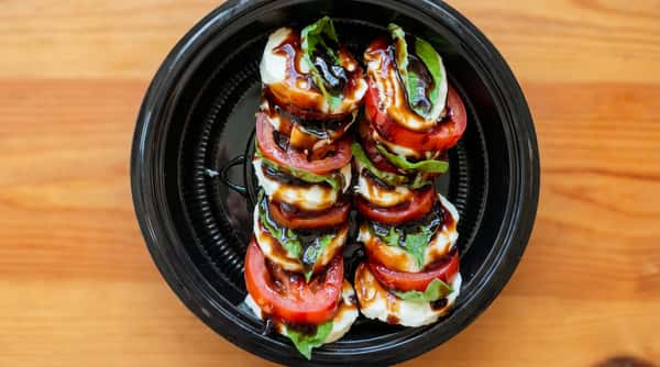Caprese salad with sliced tomatoes, fresh mozzarella, and basil drizzled with balsamic glaze in a black container.