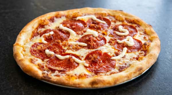 Pepperoni pizza with diced bacon and white sauce drizzles on a round crust.