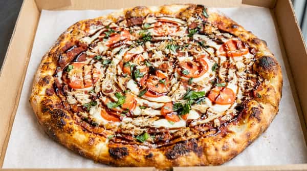 Pizza with shredded chicken, sliced tomatoes, basil, mozzarella, and a balsamic drizzle in a cardboard box.