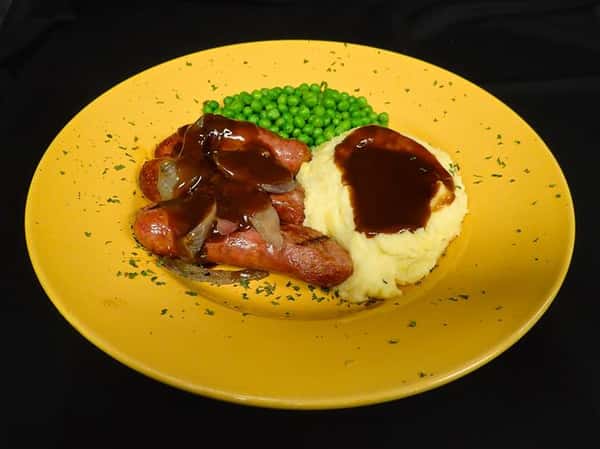 three sausages covered in onion and barbecue sauce with a side of peas and mashed potatoes with gravy on a yellow plate