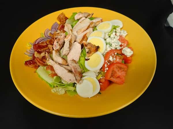 a cobb salad with boiled egg, bleu cheese crumbles, tomato, chicken, bacon, and red onion on a bed on lettuce on a yellow plate