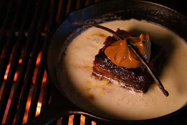 Rich and indulgent bread pudding served in a cast iron skillet over an open flame, topped with a generous dollop of caramel sauce and a vanilla bean pod, nestled in a pool of creamy custard — a warm and decadent dessert perfect for upscale comfort food or gourmet Southern cuisine settings.