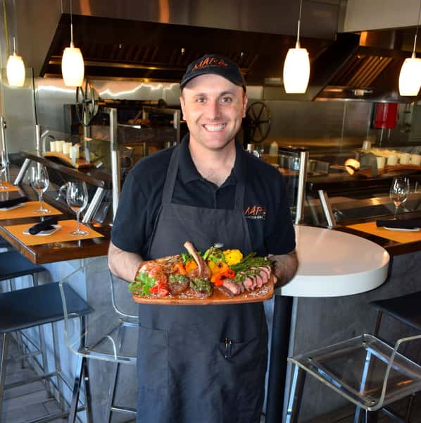 Smiling chef proudly presenting a vibrant mixed grill platter featuring lamb chops, shrimp, steak, and fire-roasted vegetables — showcasing the bold, wood-fired flavors of a modern open-kitchen restaurant.