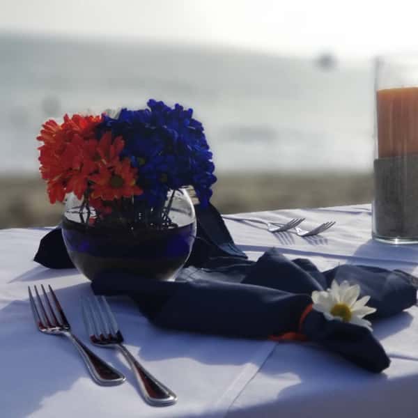 Close-up of an elegant beachfront tablescape featuring navy napkins with daisy accents, polished silverware, and a vibrant centerpiece of red and blue flowers in a glass vase — perfect for coastal weddings or sunset dinners by the sea.