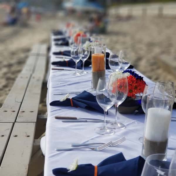 Elegant long table set directly on the beach, featuring white linens, navy napkins, candles, and vibrant floral centerpieces — a stunning setup for coastal weddings, private dinners, or luxury seaside events.