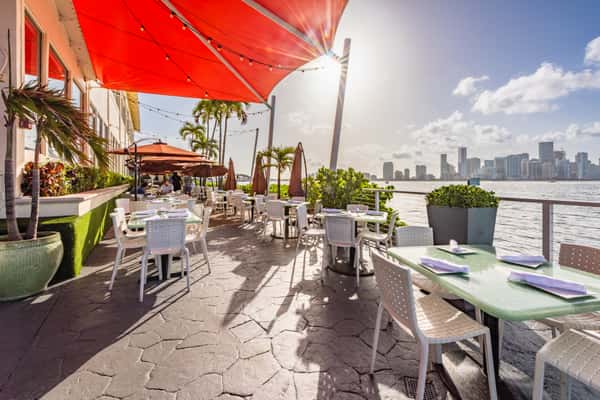 Waterfront outdoor restaurant patio with red umbrellas, marina views, and elegant open-air dining overlooking the city skyline