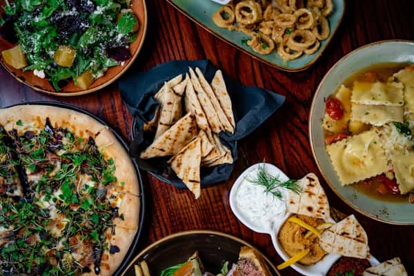Overhead view of an Italian-Mediterranean spread featuring wood-fired pizza with microgreens, grilled pita bread, spinach beet salad with goat cheese, fried calamari with lemon aioli, cheese ravioli in a tomato garlic broth, and a trio of Mediterranean dips including tzatziki and hummus on a dark wood table.